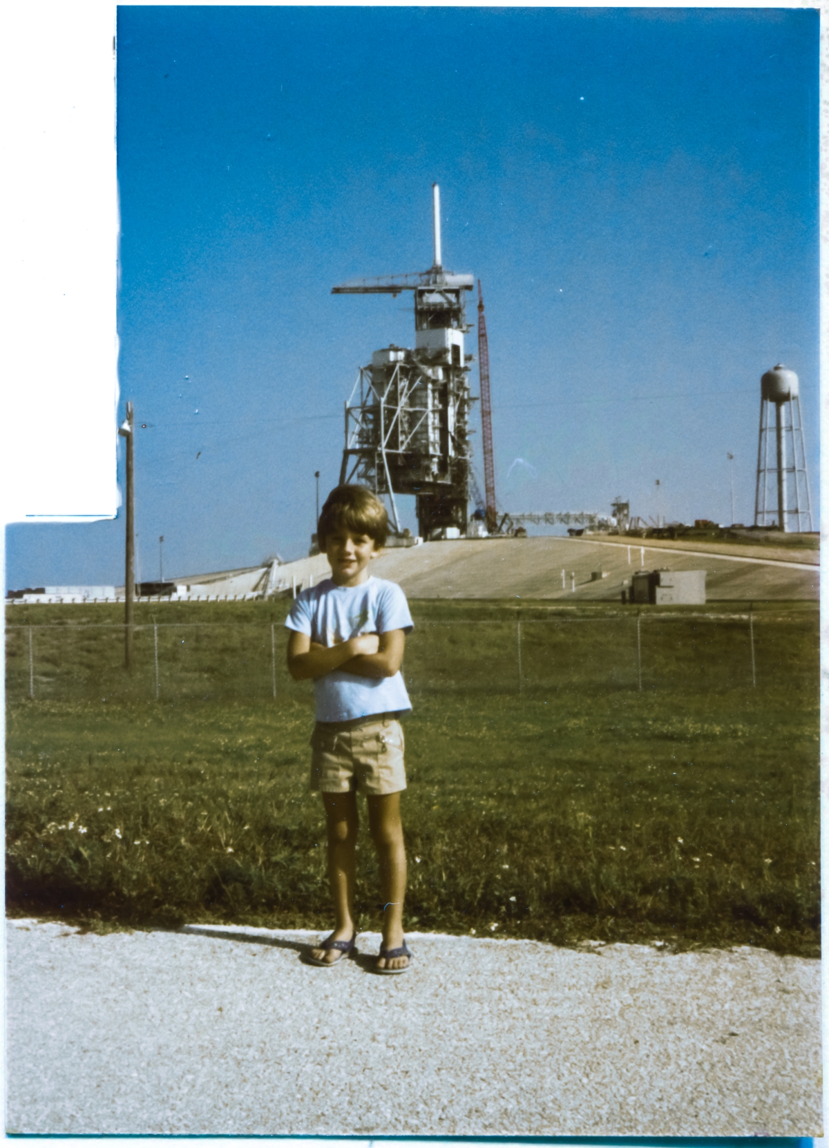 Image 085. At an age of 6 years old, an age that could not be more perfectly matched for the moment, Kai MacLaren stands on the Bypass Road just outside the Perimeter Fence of Space Shuttle Launch Complex 39-B, Kennedy Space Center, Florida. Behind him, to the northeast, the looming structures of the RSS and the FSS tower above the concrete body of the Pad, attended by large construction cranes. Kai is as much in his element as it is ever possible to be, and understands exactly how rare and sublime a moment this is, and will remember it for a lifetime. As will his father. Photo by James MacLaren.